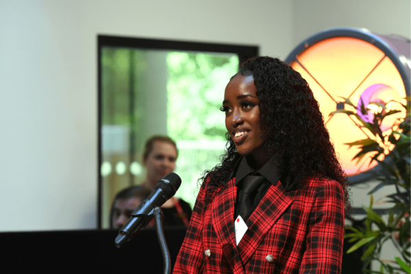 Former SCIoT student Deborah Mungai addressing the audience at the SCIoT's celebration event. Deborah is smiling at the audience and wearing a red and black checked suit.